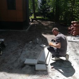 WAWER-BRUK Mariusz Wiącek - Pracownik w pomarańczowych rękawicach docina kostkę granitową na placu budowy, obok ułożone płyty, w tle budynek z brązowymi panelami i drzewa.