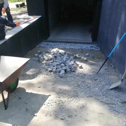 WAWER-BRUK Mariusz Wiącek - Przygotowanie podłoża pod kostkę granitową: rozsypany piasek, pryzma kostki, grabie i łopata obok wjazdu do garażu.