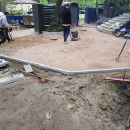 Układanie obrzeży z kostki granitowej na podsypce, zagęszczanie terenu ubijarką wibracyjną, widoczne narzędzia i materiały budowlane, w tle kontener i schody.