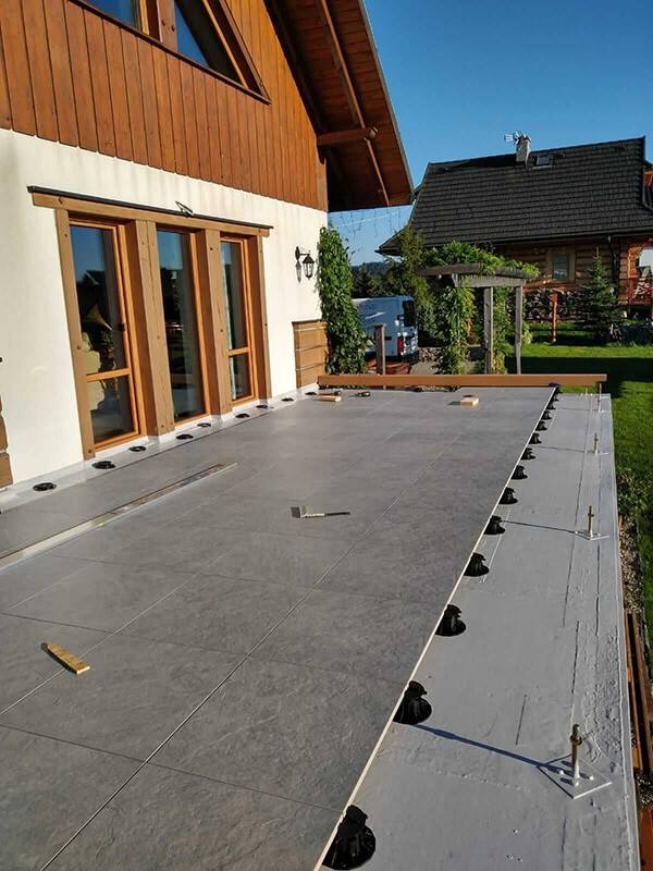 Montaż tarasu wentylowanego z szarych płyt na regulowanych wspornikach, widoczne elementy konstrukcyjne i poziomice, w tle budynek z drewnianymi elementami.