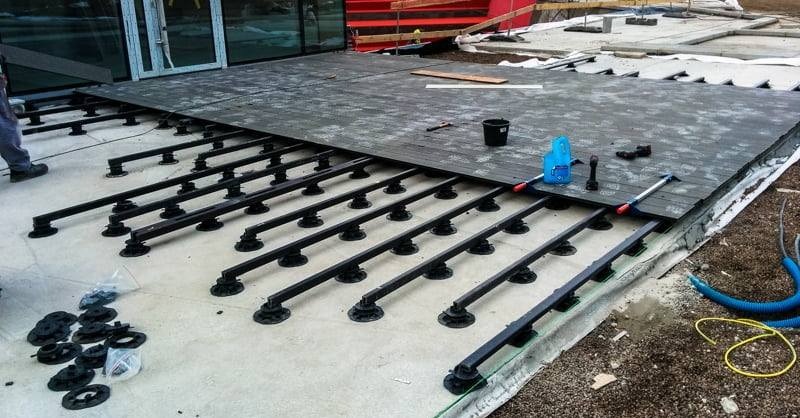 Montaż tarasu wentylowanego: widok na konstrukcję wsporczą z regulowanymi wspornikami i częściowo ułożone deski kompozytowe, narzędzia pracy i wiadro w tle.