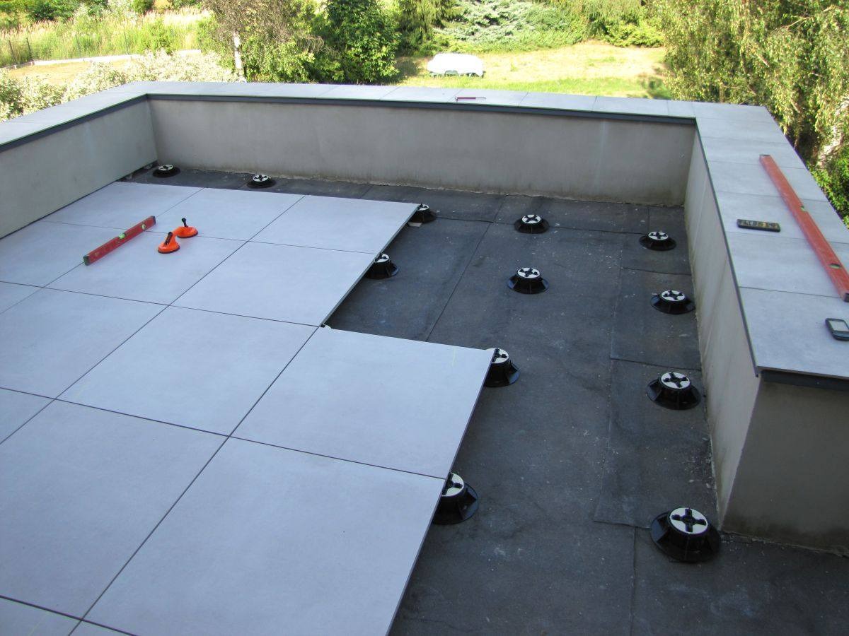 Montaż tarasu wentylowanego: jasnoszare płytki układane na regulowanych wspornikach, poziomica i przyssawki do płytek obok, widok z góry na częściowo wykończony taras.