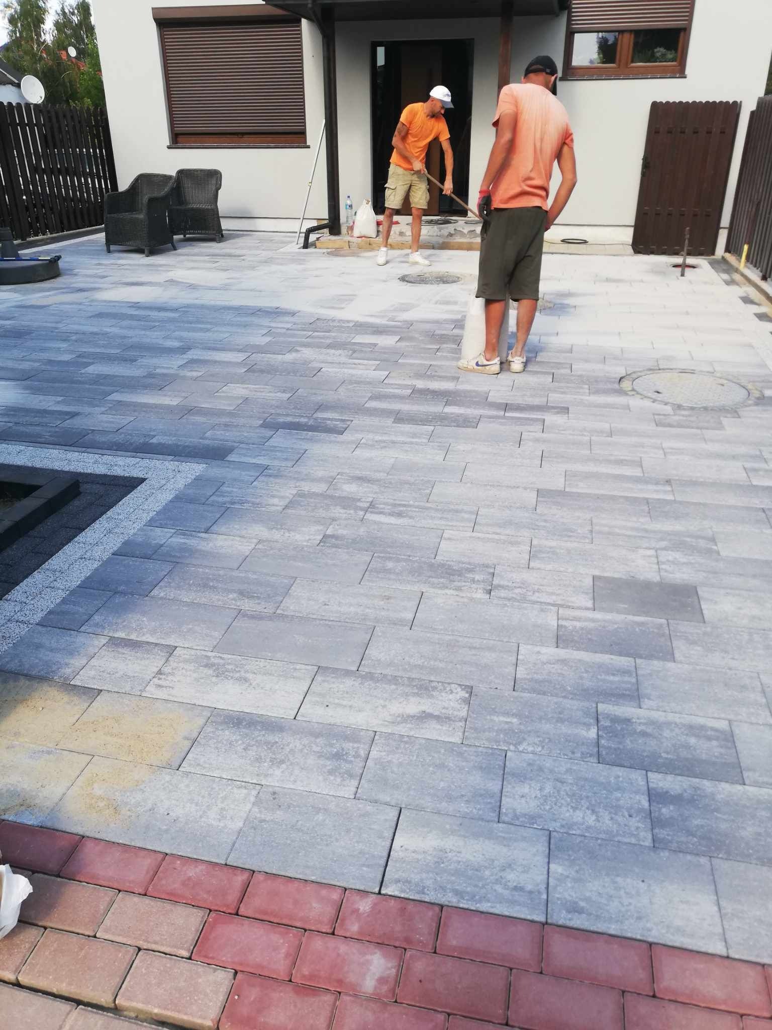 Dwóch pracowników wyrównuje powierzchnię podłoża pod szarą kostkę brukową na posesji z elementami czerwonej kostki, widoczne narzędzia i materiały budowlane.