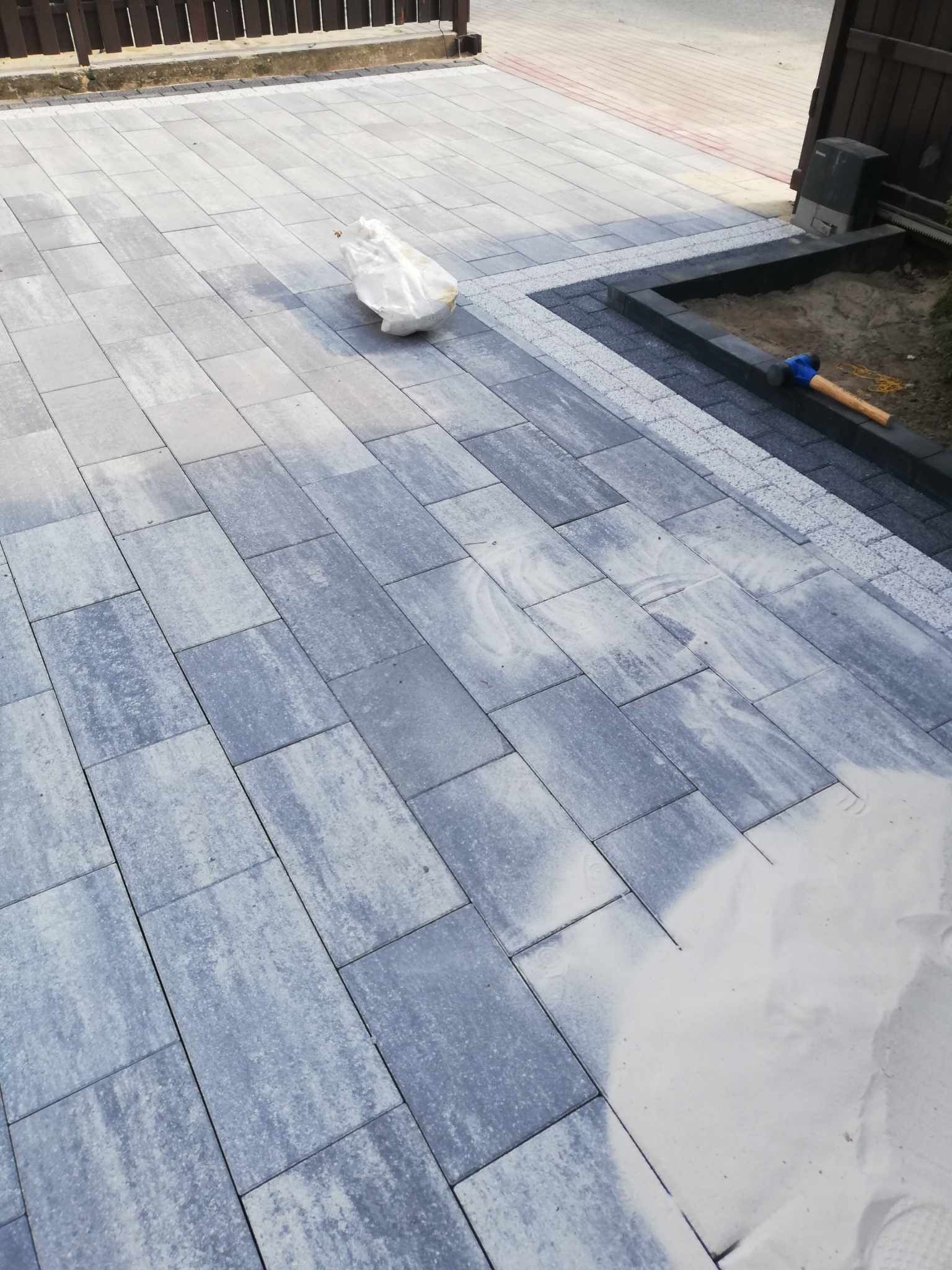 Świeżo ułożona szara kostka brukowa o nieregularnym wzorze z widocznym workiem z piaskiem do zasypywania fug, fragment bramy w tle i gumowy młotek przy obrzeżu.