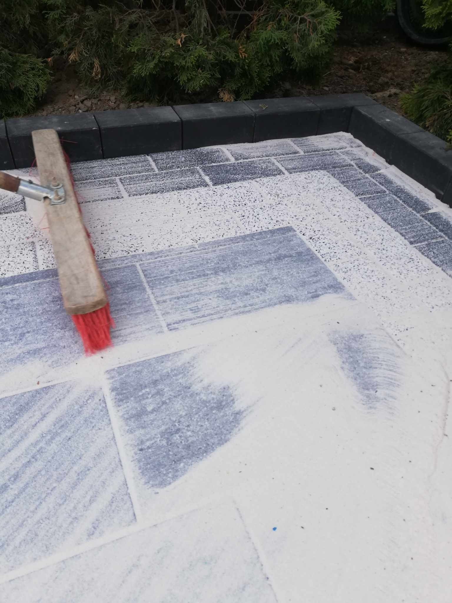 Szczotka z czerwonym włosiem rozprowadza biały piasek kwarcowy po ułożonej szarej kostce brukowej z czarnym obramowaniem, widok z góry na fragment posesji.