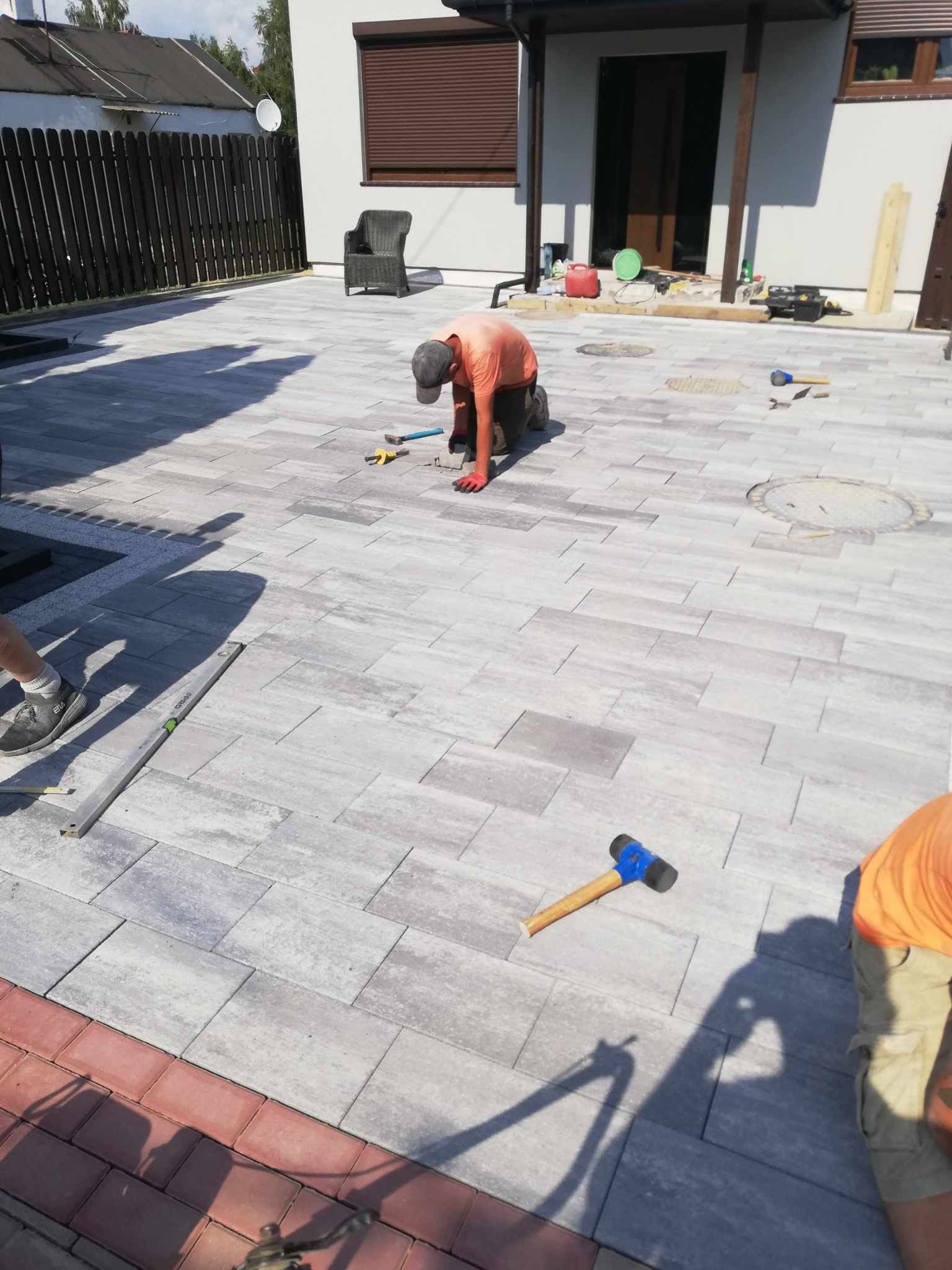 Pracownik w pomarańczowej koszulce i rękawicach układa szare kostki brukowe na podjeździe, widoczny gumowy młotek z niebieską główką, poziomica i fragment czerwonej obwódki z kostki.