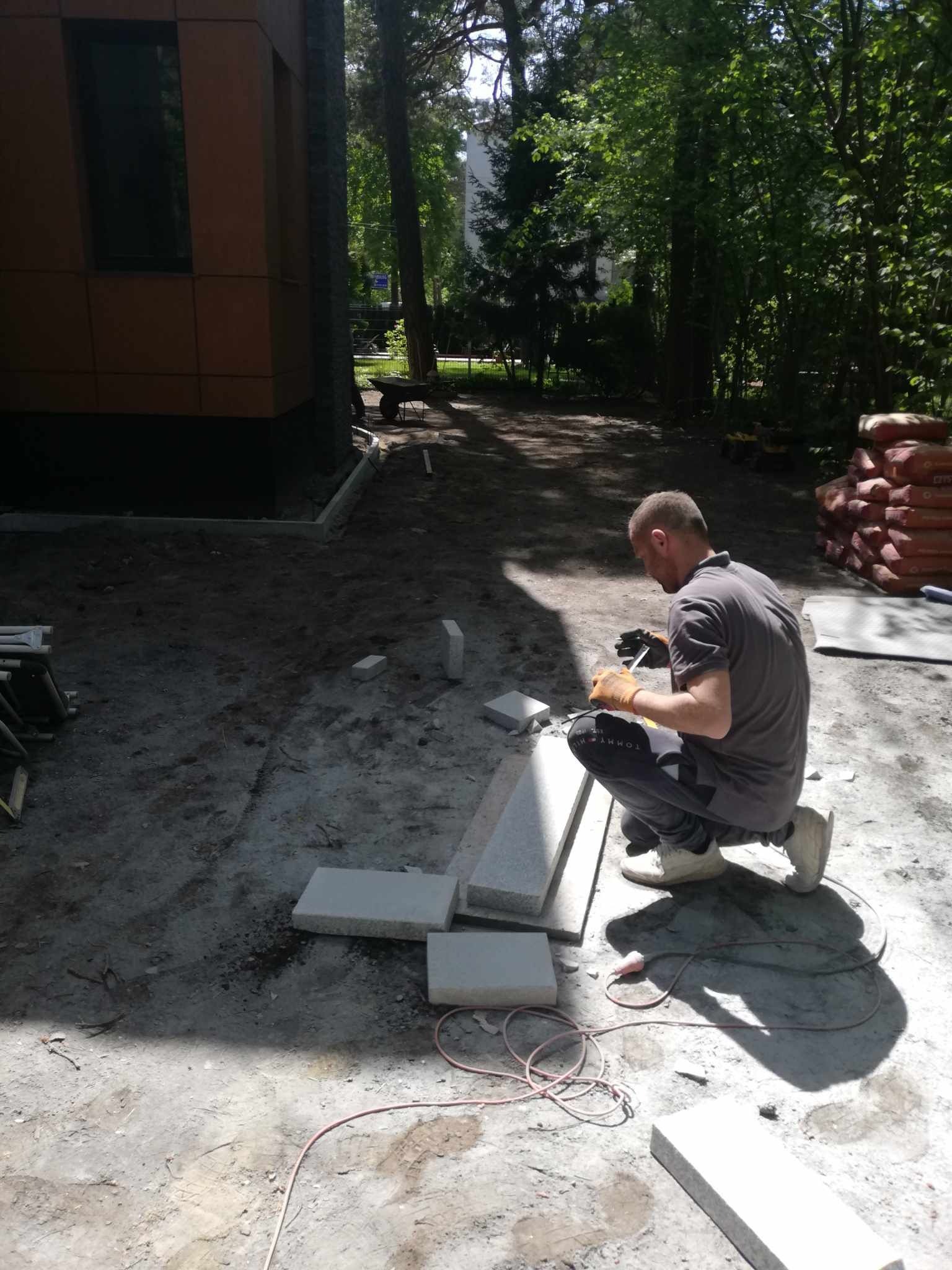 Pracownik w pomarańczowych rękawicach docina kostkę granitową na placu budowy, obok ułożone płyty, w tle budynek z brązowymi panelami i drzewa.