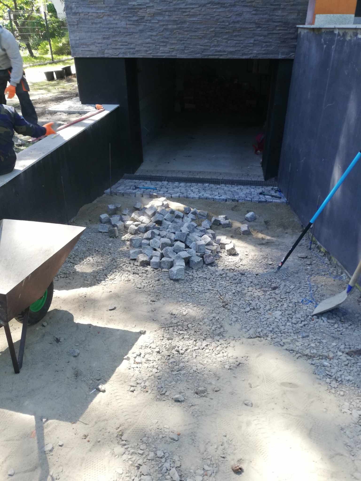 Przygotowanie podłoża pod kostkę granitową: rozsypany piasek, pryzma kostki, grabie i łopata obok wjazdu do garażu.