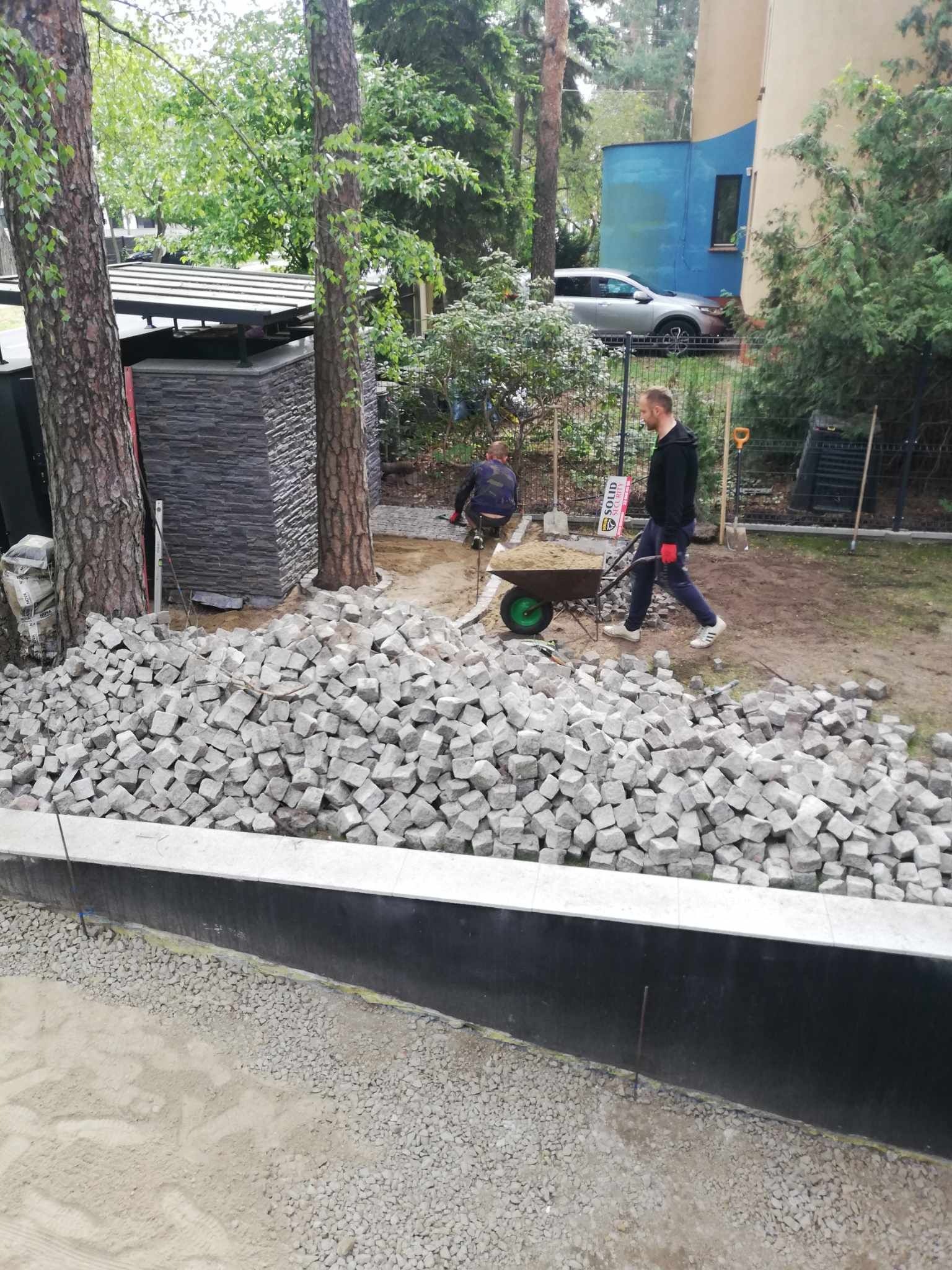 Stos szarej kostki granitowej na placu budowy, robotnik z taczką w tle, widoczne narzędzia i przygotowane podłoże.