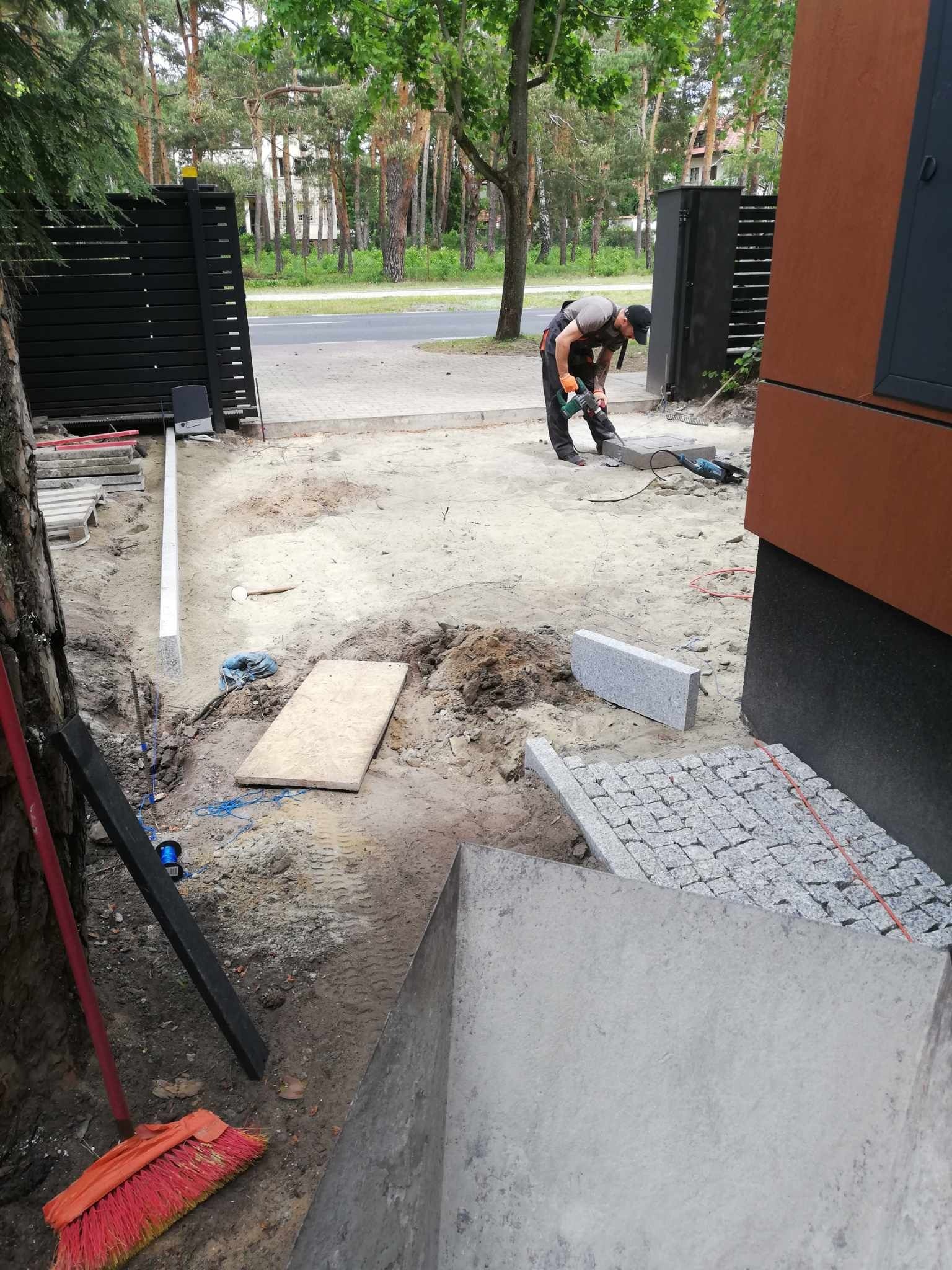 Pracownik przycina granitową kostkę brukową szlifierką kątową na posesji z nowoczesnym ogrodzeniem i budynkiem, przygotowując nawierzchnię w Radosci.