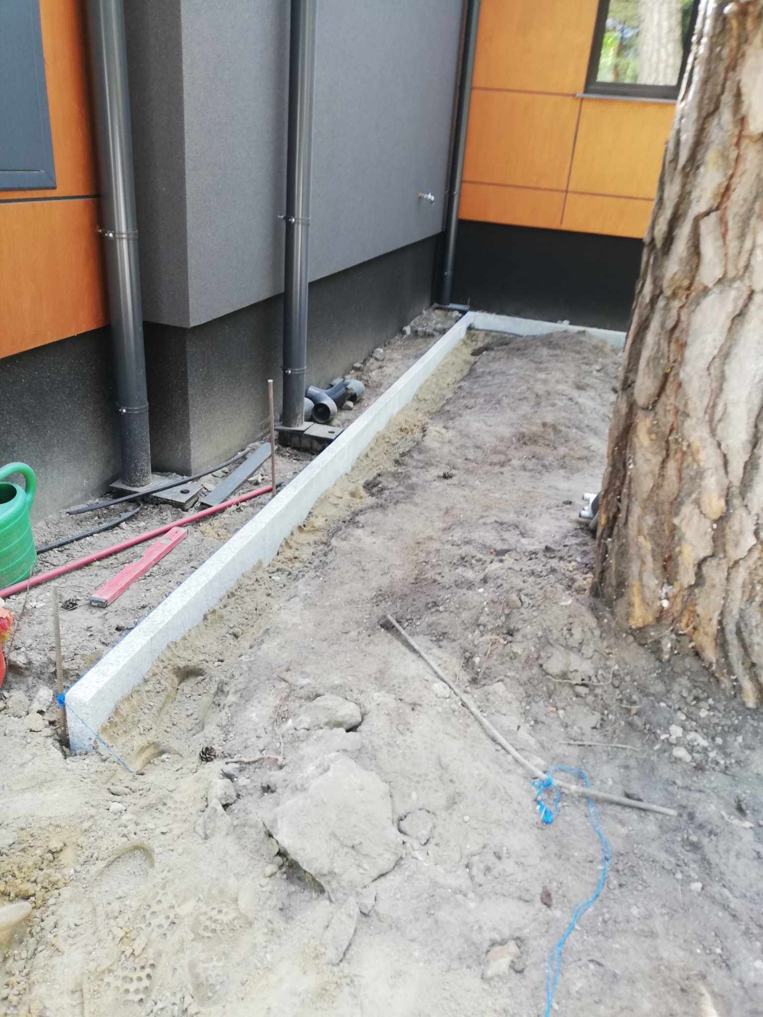 Montaż obrzeża z jasnego kamienia wzdłuż ściany budynku z widocznymi narzędziami i elementami instalacji, ziemia wokół przygotowana pod dalsze prace.