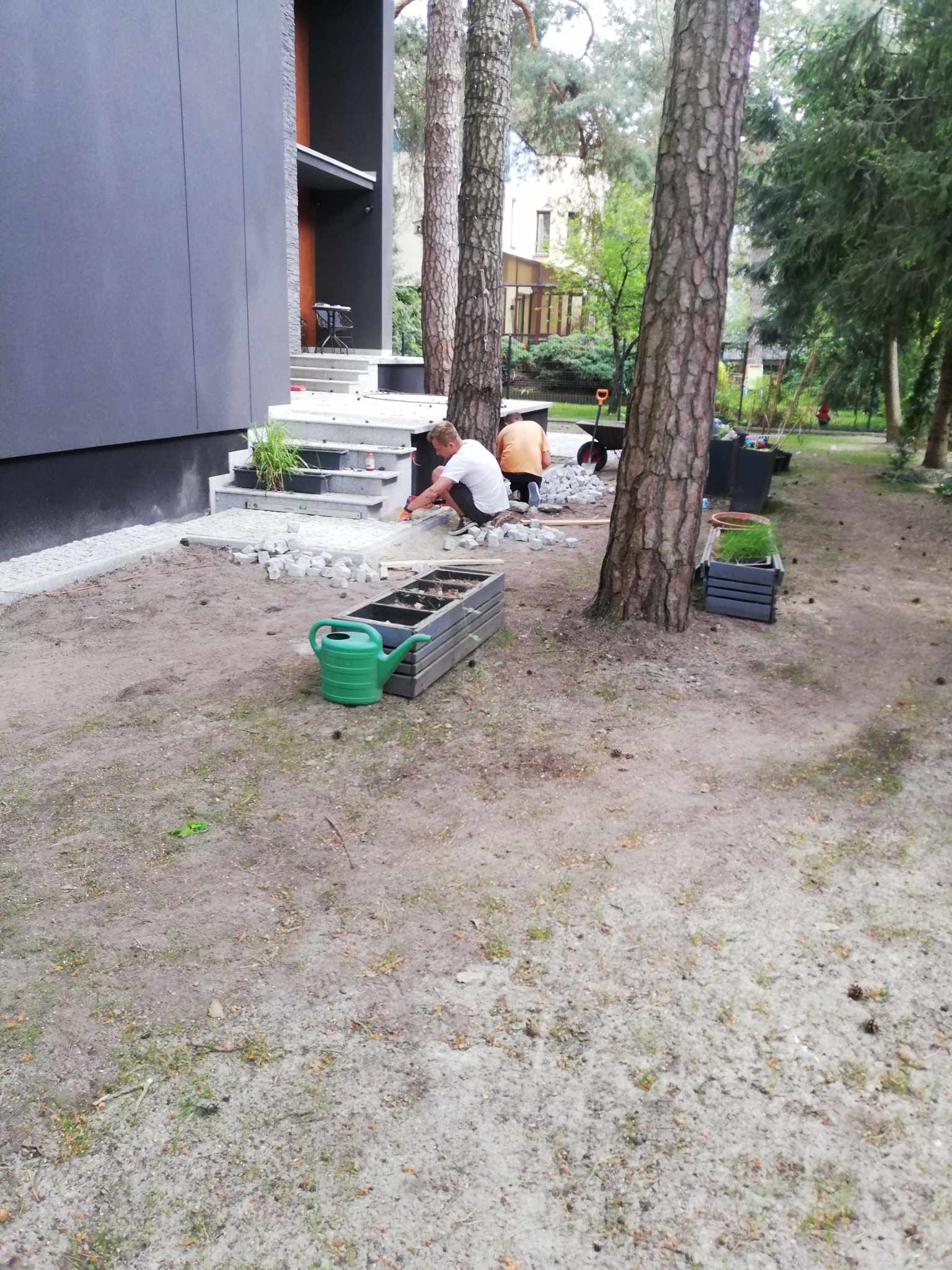 Prace brukarskie w ogrodzie: dwóch mężczyzn układa kostkę granitową przy schodach nowoczesnego domu z szarą elewacją, obok leży zielona konewka i paleta z kostką, w tle drzewa.