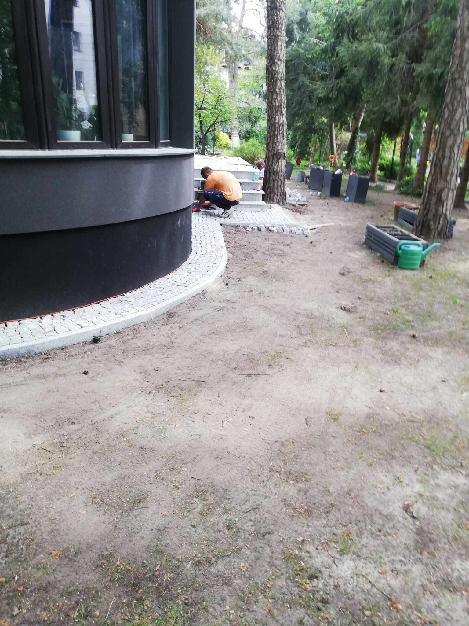 Pracownik w pomarańczowej koszulce układa kostkę granitową wokół zaokrąglonej ściany budynku, widoczne narzędzia, w tle drzewa i donice.