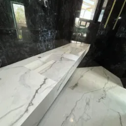 Nowoczesna łazienka z czarnymi, błyszczącymi ścianami i długim, białym blatem z marmurowym wzorem, w którym wbudowane są dwie umywalki. Podłoga wyłożona jest płytkami w podobnym marmurowym wzorze.