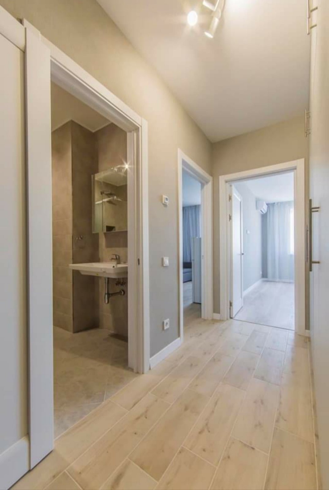 Korytarz z drewnopodobnymi płytkami podłogowymi, widok na łazienkę z umywalką na stelażu i lustrem oraz pokój z klimatyzacją i zasłonami.