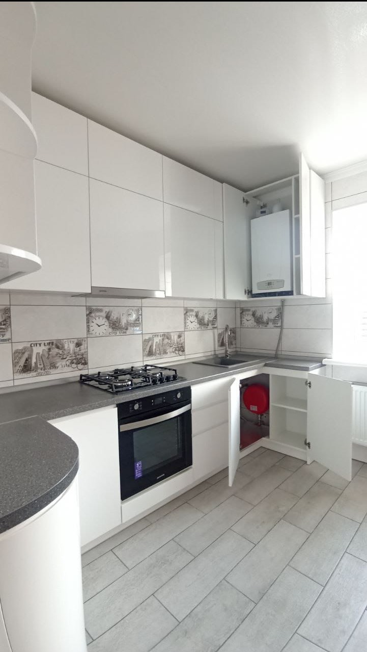 Nowoczesna, jasna kuchnia z białymi, błyszczącymi szafkami, szarym blatem i płytkami z motywem miejskim nad blatem. Widoczny piekarnik, płyta gazowa, zlew oraz czerwony zbiornik w szafce pod zlewem...