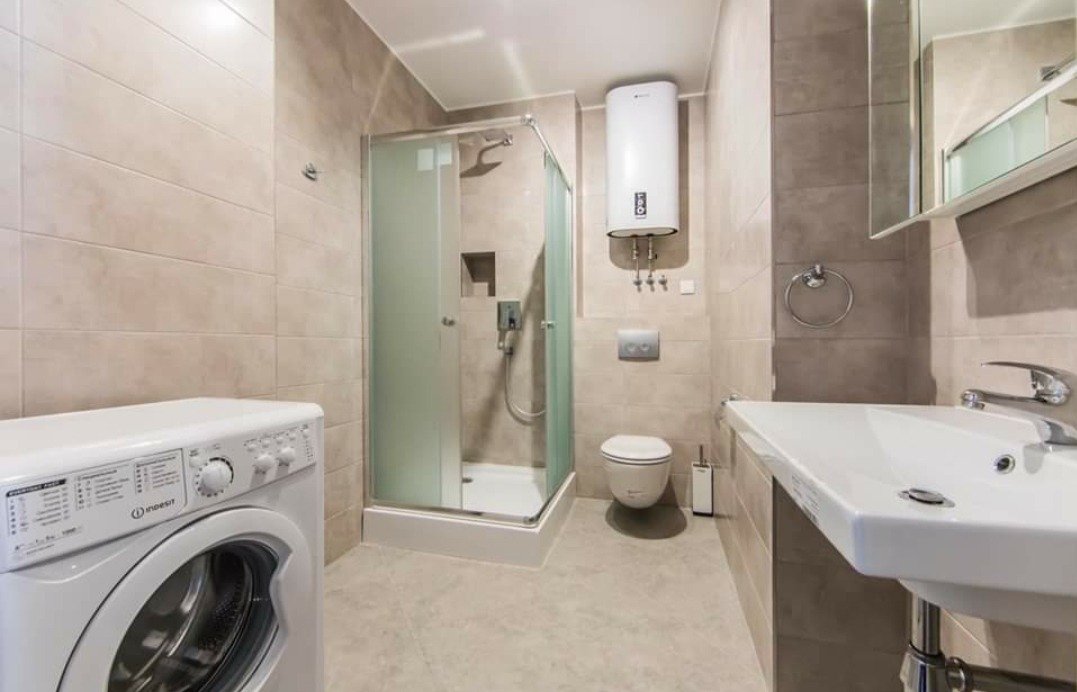 Jasna łazienka z kabiną prysznicową, pralką Indesit i grzejnikiem elektrycznym, wykończona płytkami w odcieniach beżu i szarości.