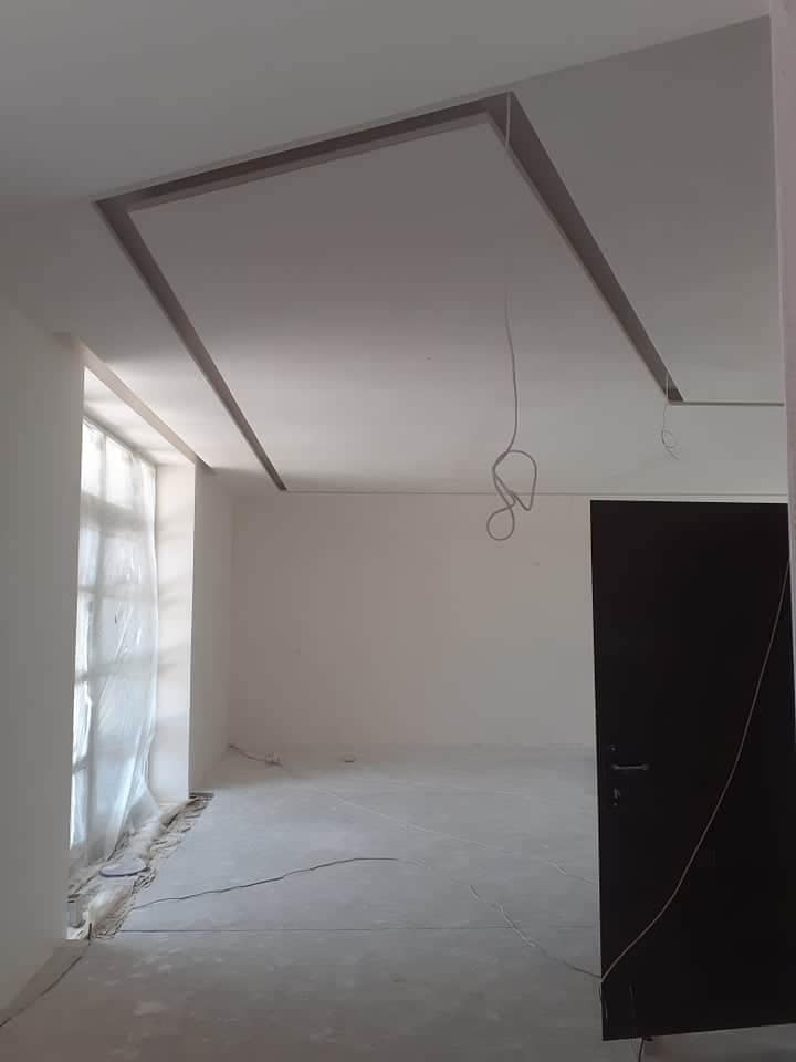 Wnętrze pokoju w trakcie remontu, widoczne świeżo pomalowane ściany i sufit z dekoracyjną ramą, podłoga przygotowana do dalszych prac, okno zasłonięte folią malarską, wiszące kable elektryczne.