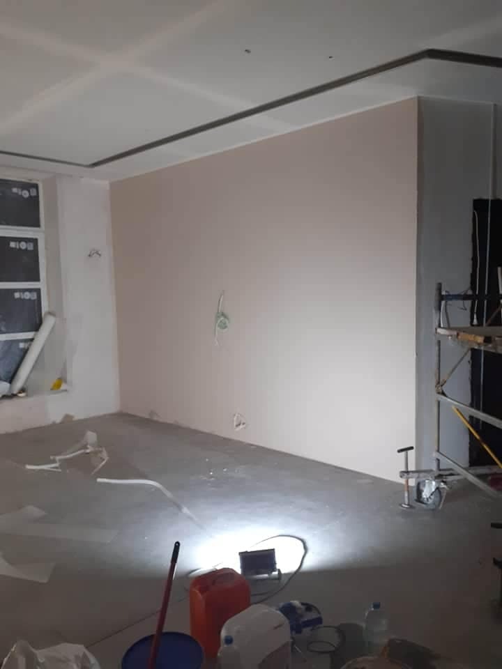 Wnętrze w trakcie prac wykończeniowych: ściana po szpachlowaniu na gładko, z widocznymi puszkami elektrycznymi, sufit z płyt gipsowo-kartonowych z dekoracyjną listwą, na podłodze narzędzia...