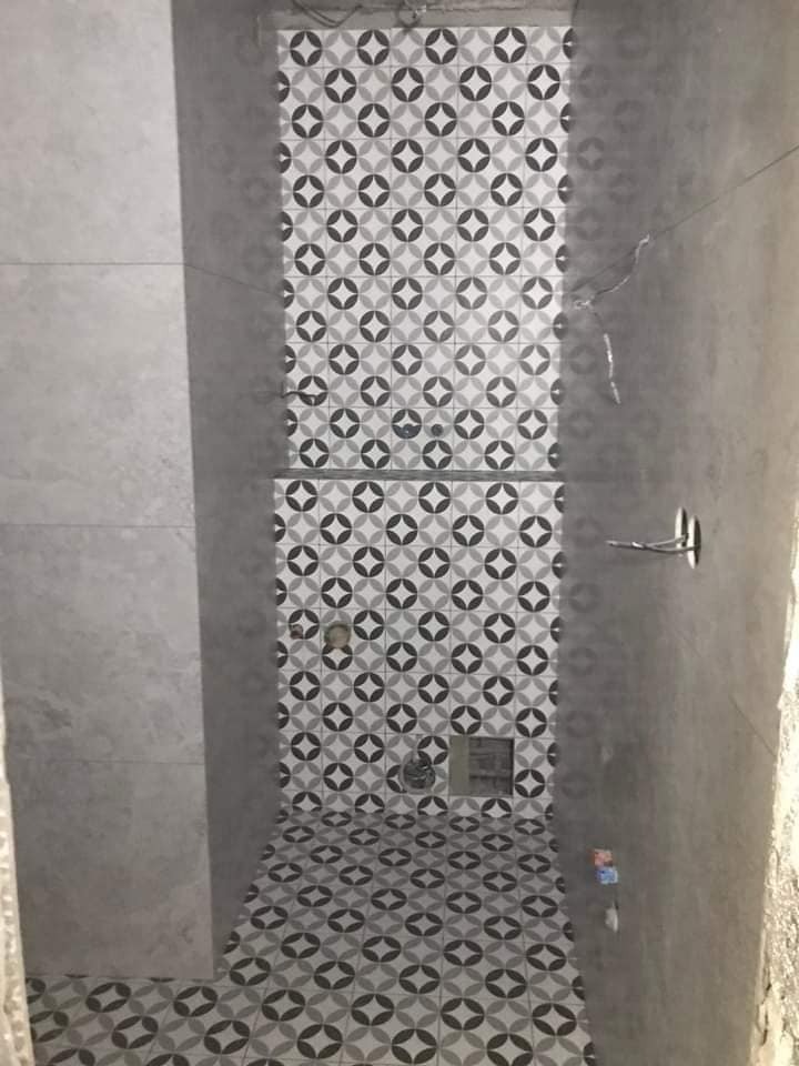 Wnętrze łazienki w trakcie remontu, z ułożoną wzorzystą glazurą w odcieniach szarości na podłodze i ścianach, widoczne niedokończone ściany i instalacje.