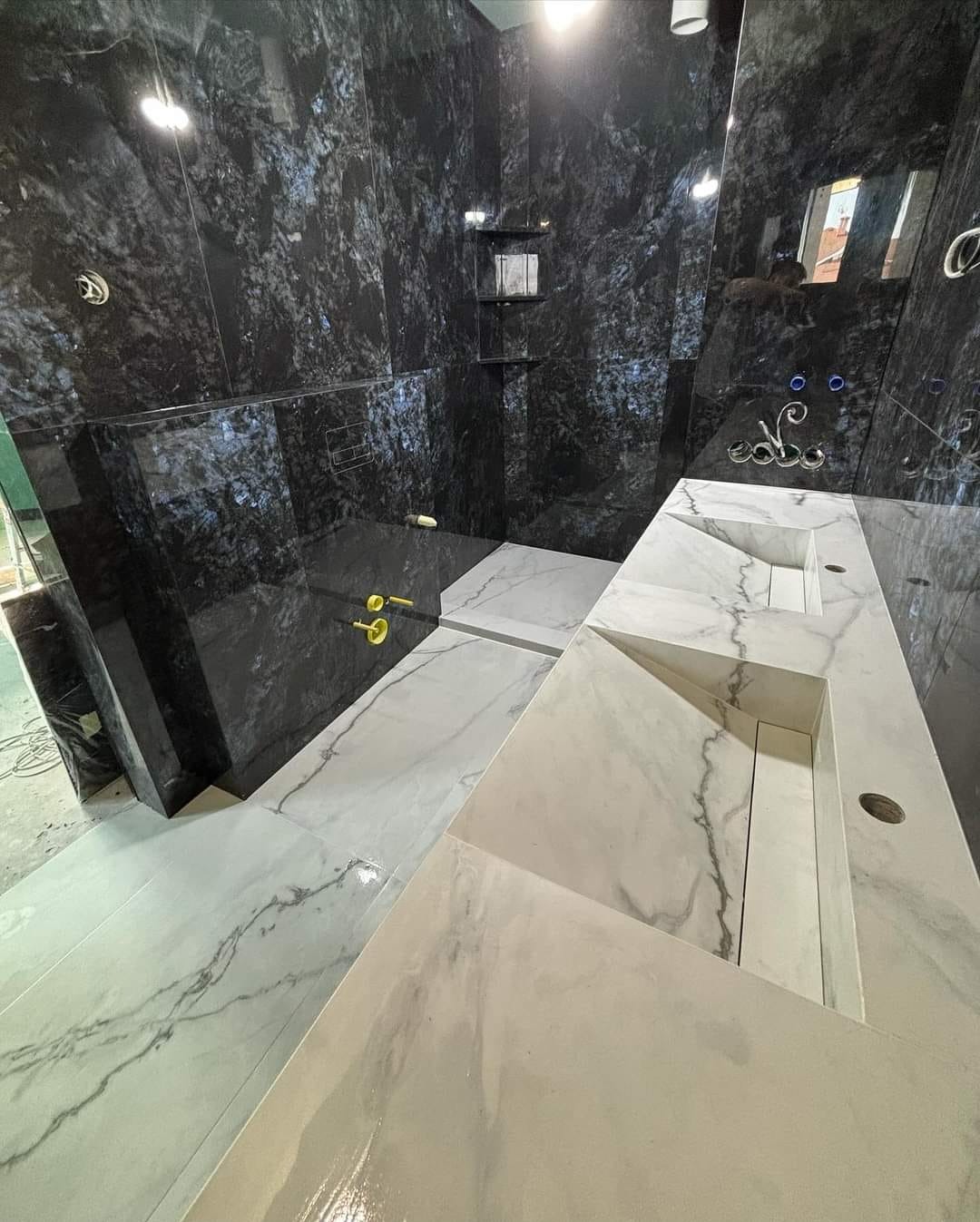 Nowoczesna łazienka z czarnymi ścianami z płytek imitujących kamień i białymi marmurowymi umywalkami w geometrycznym stylu, złote baterie, oświetlenie sufitowe.