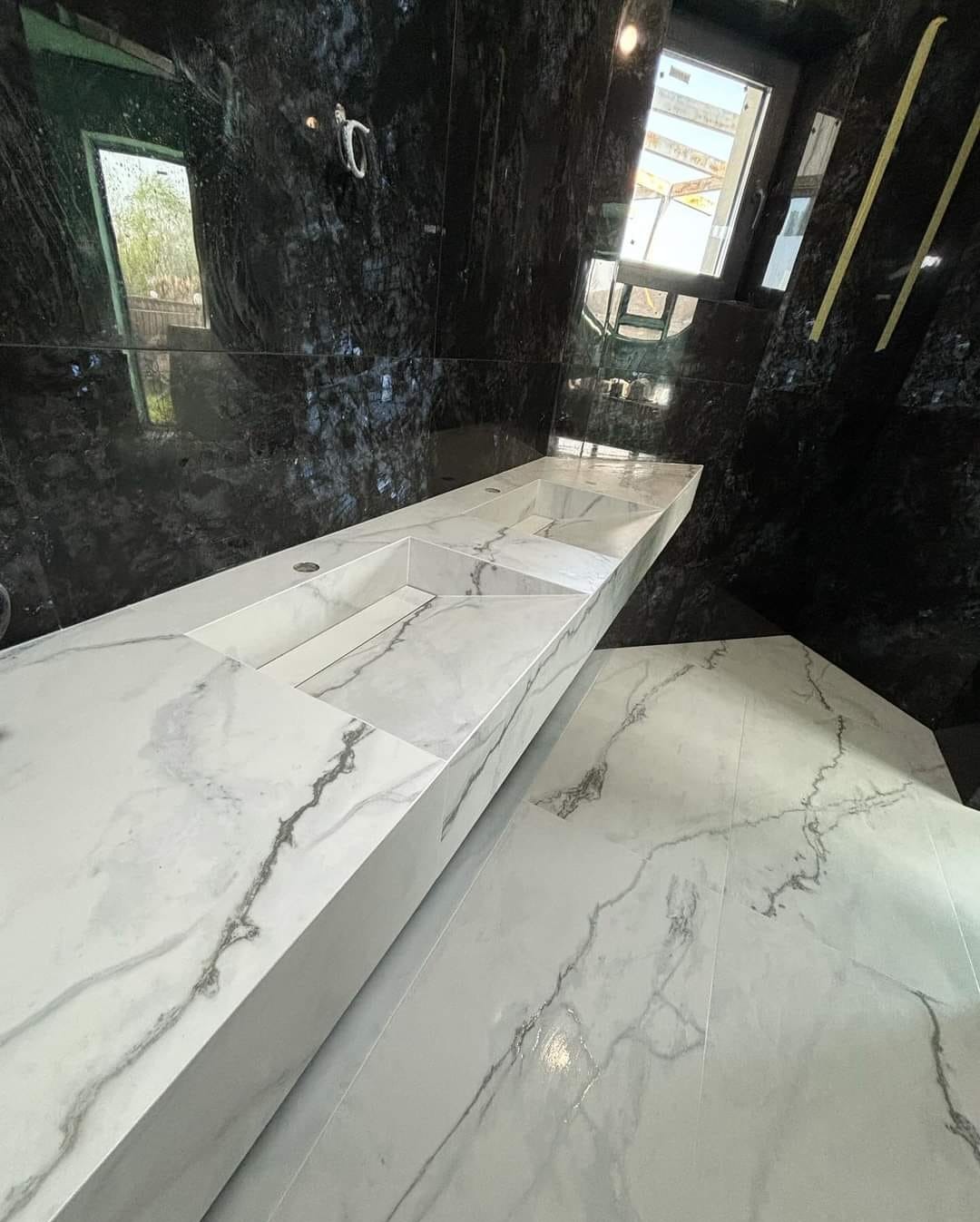 Nowoczesna łazienka z czarnymi, błyszczącymi ścianami i długim, białym blatem z marmurowym wzorem, w którym wbudowane są dwie umywalki. Podłoga wyłożona jest płytkami w podobnym marmurowym wzorze.