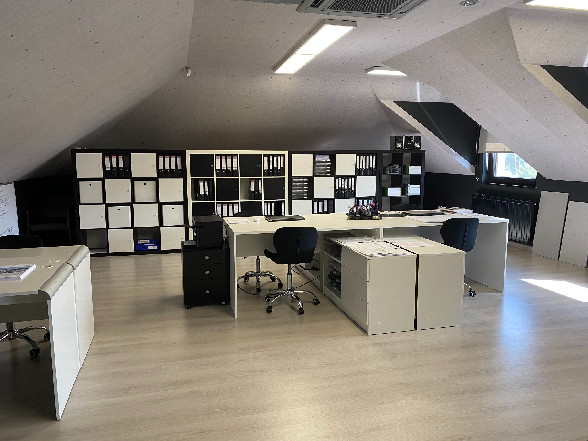 Przestronne biuro w adaptowanym poddaszu z białymi biurkami, czarnymi krzesłami obrotowymi i regałem z segregatorami, oświetlone panelami LED.