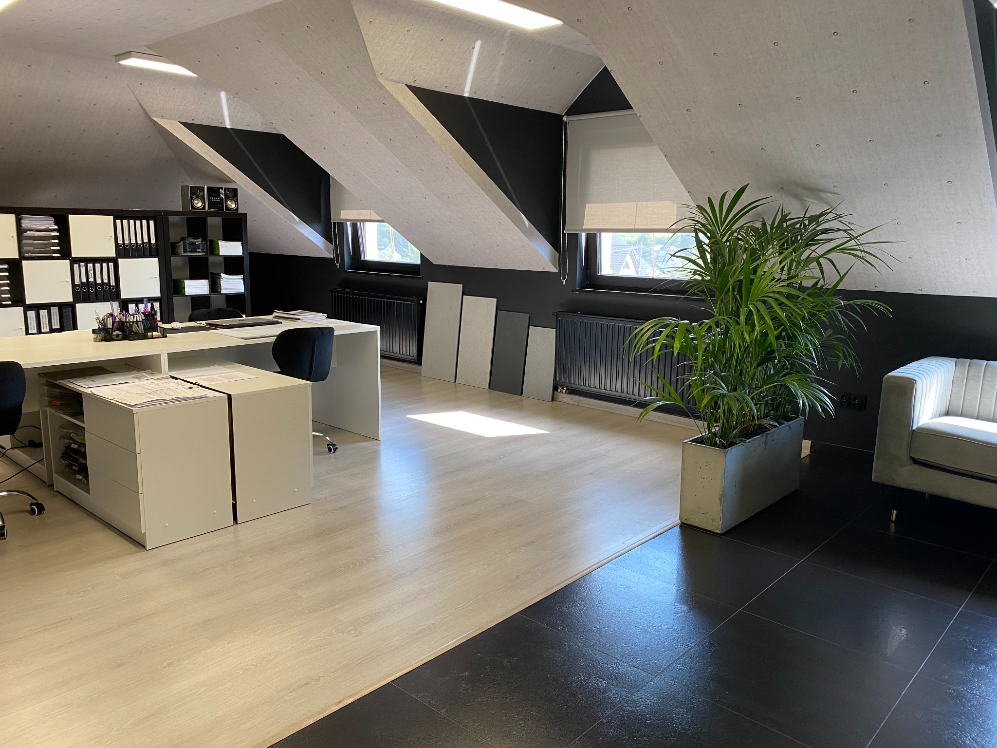 Jasne biuro na poddaszu z biurkiem, szafkami, regałem na dokumenty, rośliną doniczkową i szarą sofą; charakterystyczne skośne sufity z elementami wykończeniowymi w kolorze czarnym.