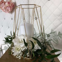 Projekt Event - Dekoracja stołu z geometrycznym, złotym lampionem, białą świecą i kompozycją z białych kwiatów oraz zielonych liści na tle białej, pikowanej ściany.