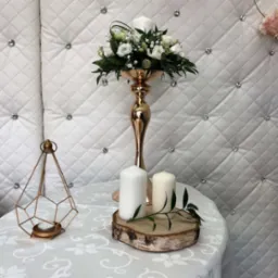 Dekoracja stołu z wysokim, złotym świecznikiem, białymi kwiatami, świecami na drewnianym plastrze i metalowym lampionem na tle pikowanego panelu z kryształkami.