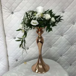 Kompozycja kwiatowa z białych róż i gipsówki w pozłacanym wazonie na stole, tło stanowi pikowana ściana z dekoracyjnymi kryształkami.