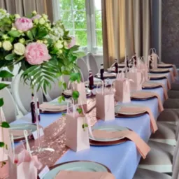 Elegancko nakryty stół weselny z kwiatową dekoracją, różowym bieżnikiem z cekinami, upominkami dla gości w różowych torebkach i butelkami z napojami.