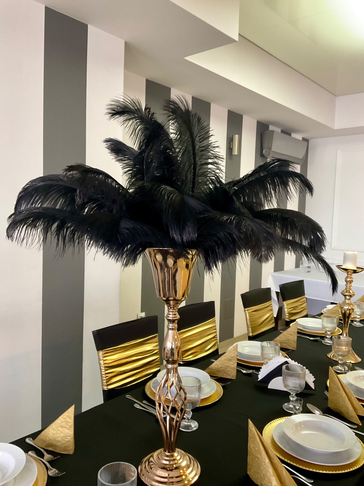 Elegancka dekoracja stołu z wysokim, złotym wazonem wypełnionym czarnymi piórami, otoczona zastawą w kolorach złota i bieli, na czarnym obrusie, z krzesłami ozdobionymi złotymi szarfami.
