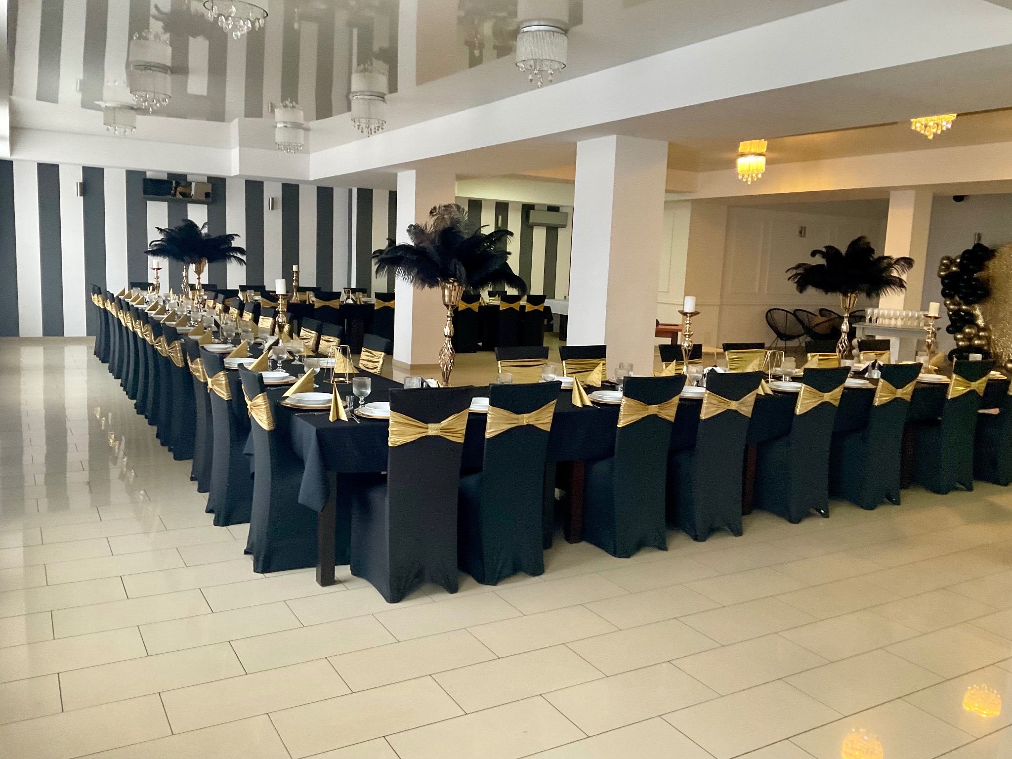 Uroczysta sala bankietowa z długim stołem w kształcie litery U, nakrytym czarnym obrusem, ze złotymi dekoracjami na krzesłach i stołach, przygotowana na przyjęcie.