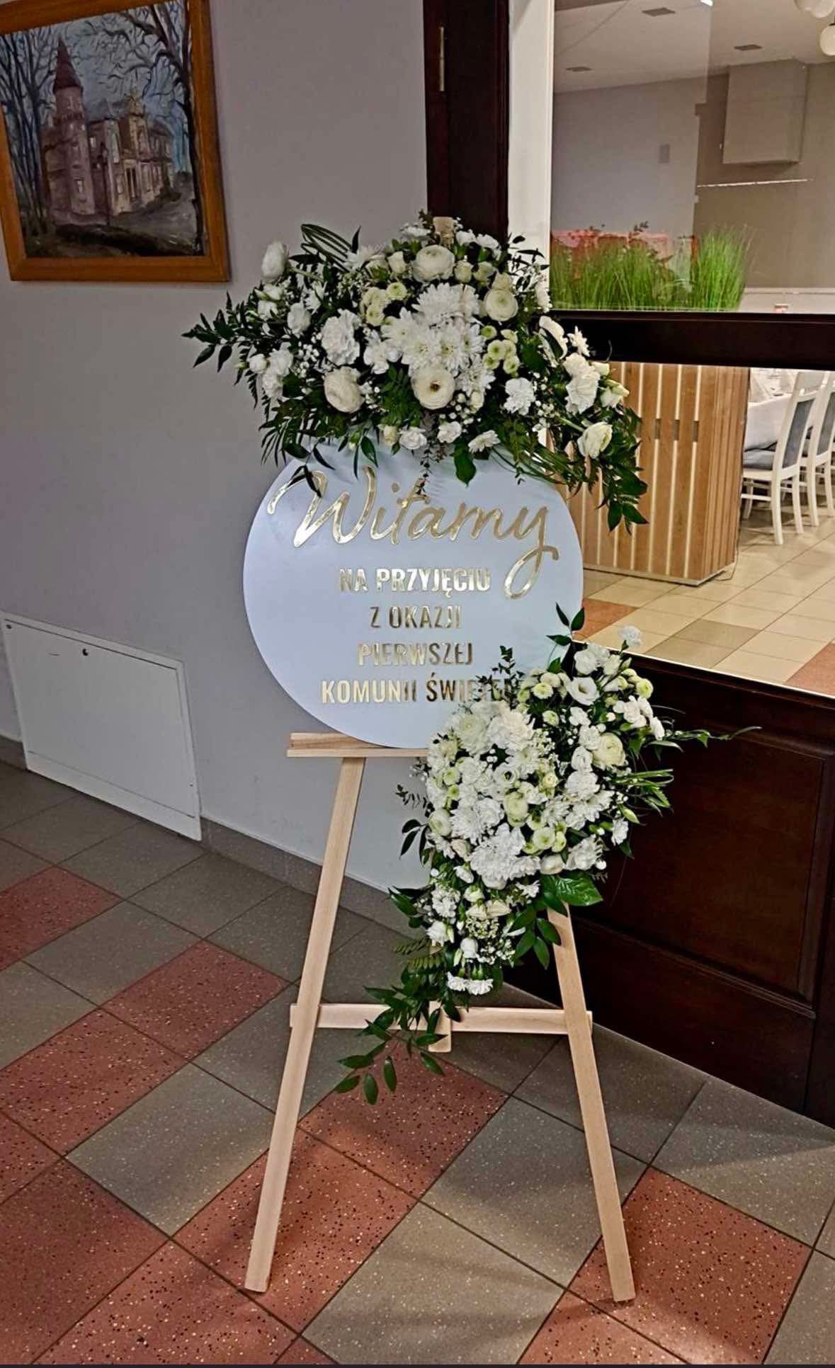 Dekoracja sali na przyjęcie komunijne: okrągła tablica powitalna z napisem 'Witamy na przyjęciu z okazji Pierwszej Komunii Świętej' w złotym kolorze, ozdobiona kompozycjami z białych kwiatów...
