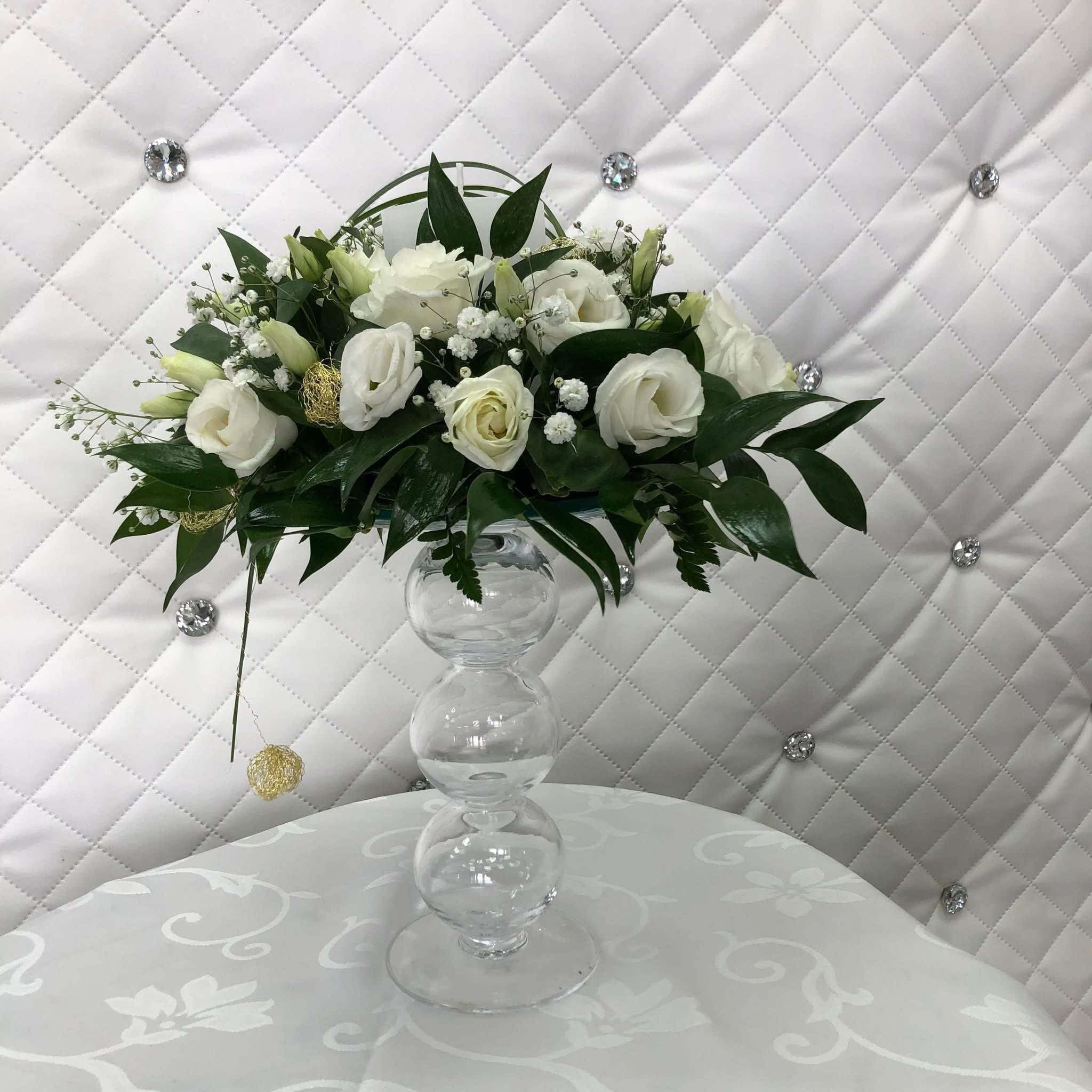 Elegancka kompozycja kwiatowa z białych róż i delikatnych dodatków w szklanym wazonie na stole z białym obrusem w kwiatowe wzory, na tle białej, pikowanej ściany z kryształkami