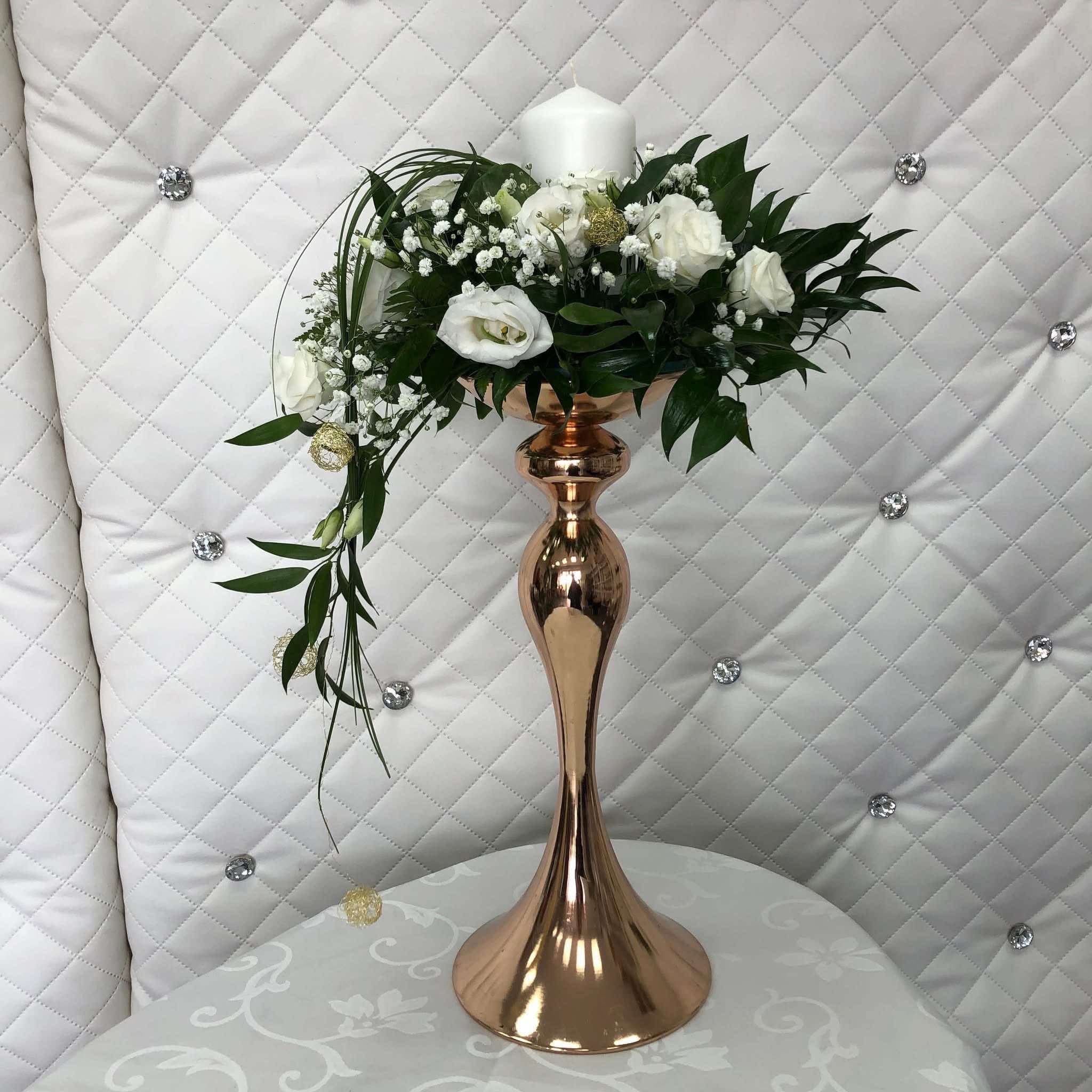 Kompozycja kwiatowa z białych róż i gipsówki w pozłacanym wazonie na stole, tło stanowi pikowana ściana z dekoracyjnymi kryształkami.