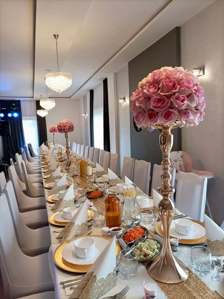 Długi stół bankietowy z białymi krzesłami, nakryty złotymi podkładkami, białymi talerzami, serwetkami z koronkowym brzegiem i wysokimi złotymi świecznikami z bukietami różowych róż, oświetlony...