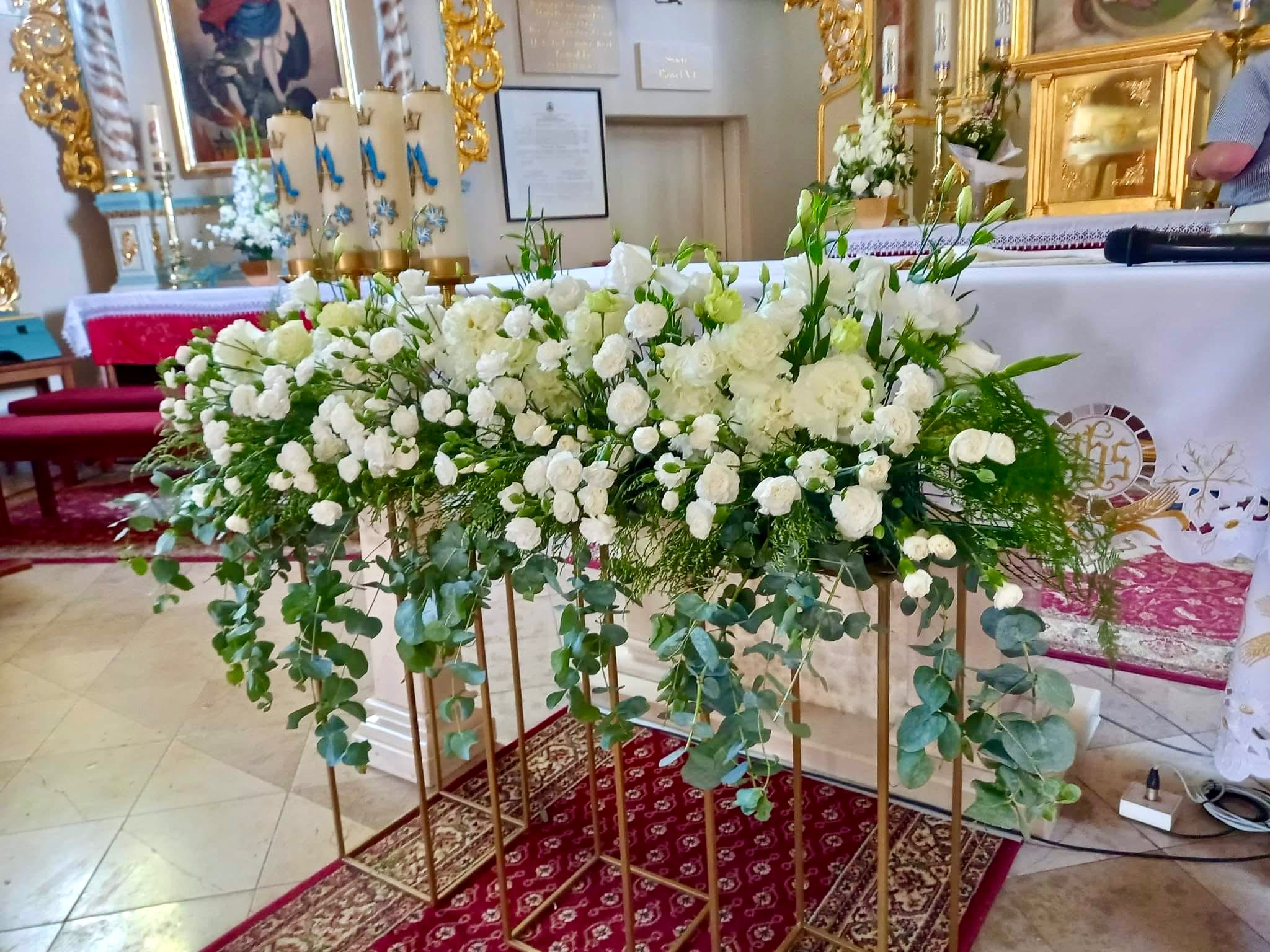 Dekoracja kwiatowa z białych róż i zielonych liści eukaliptusa na złotych stojakach, ustawiona w kościele przed ołtarzem.