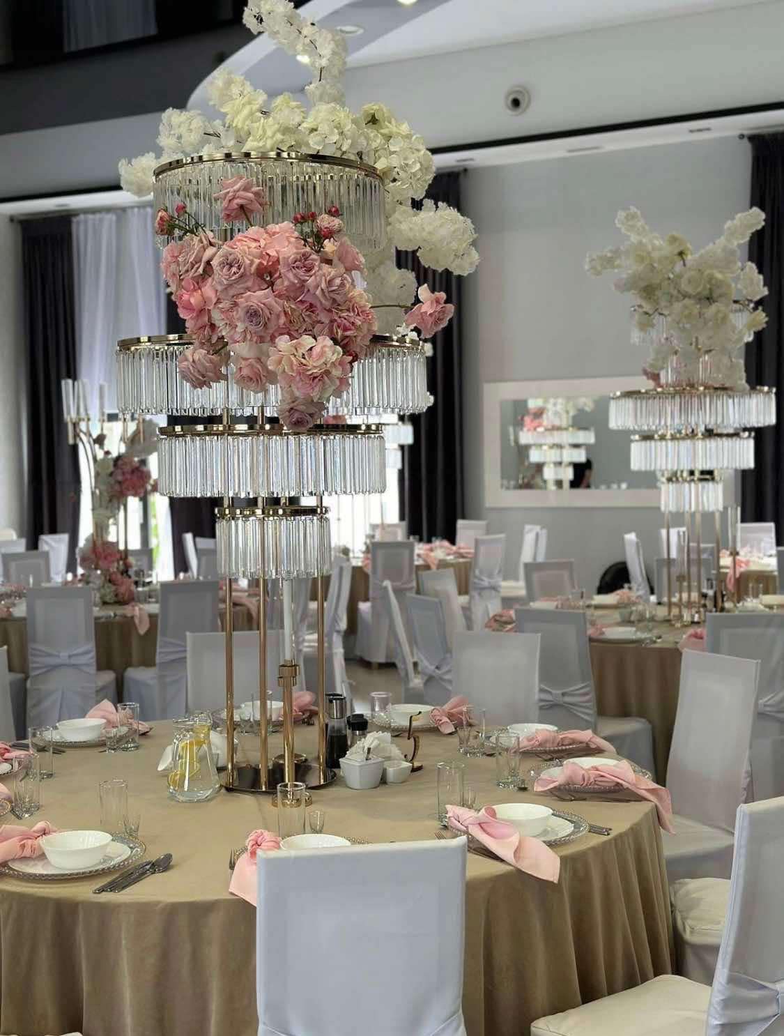 Uroczysta sala bankietowa z okrągłymi stołami nakrytymi beżowymi obrusami, białymi krzesłami z kokardami i wysokimi, wielopoziomowymi dekoracjami kwiatowymi z kryształkami.