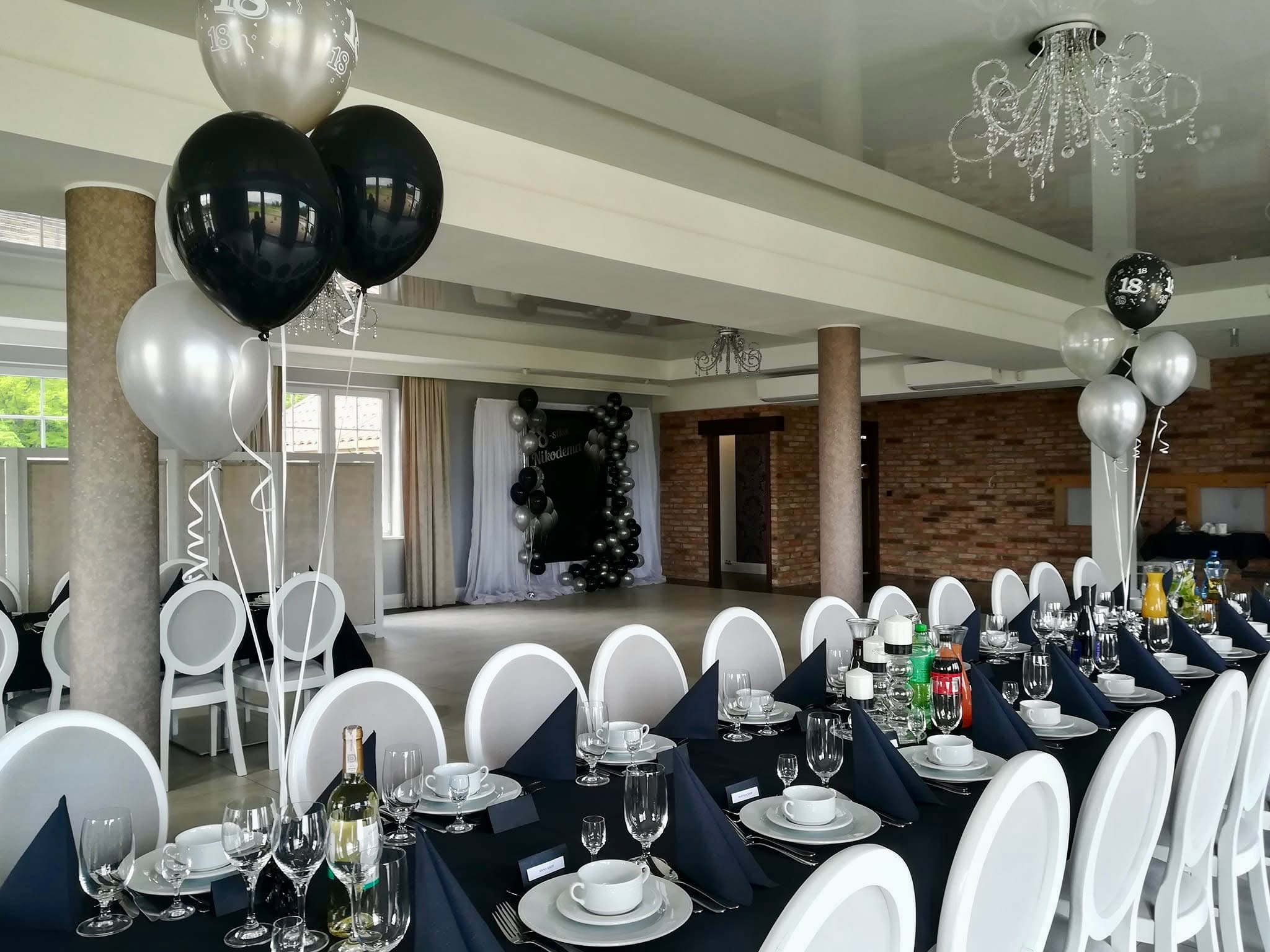 Uroczysty stół nakryty czarnym obrusem, biała zastawa, dekoracje z czarno-srebrnych balonów, przygotowany na przyjęcie w przestronnej sali z kolumnami i żyrandolem.