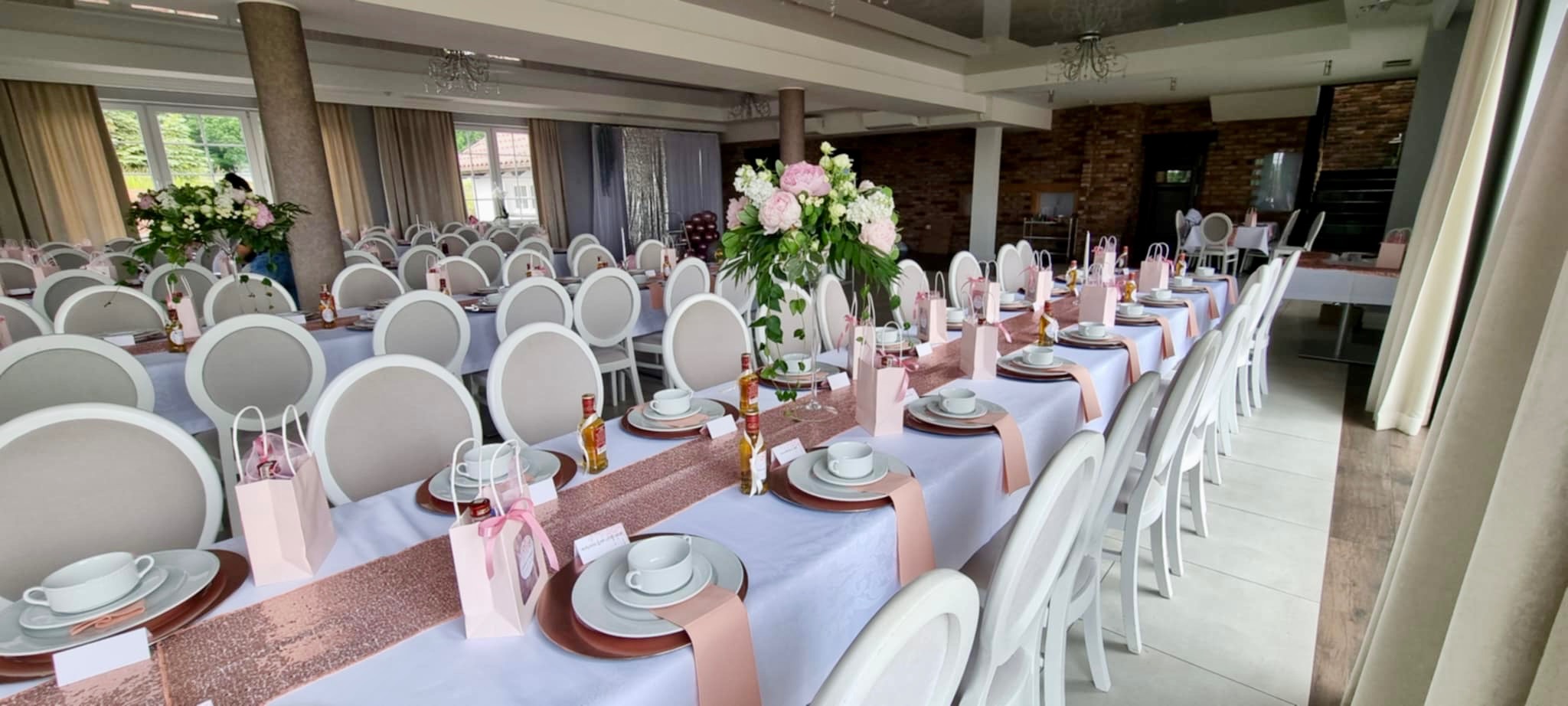 Uroczysta sala bankietowa z długim stołem, nakrytym białą tkaniną i różowo-złotym bieżnikiem, z białymi krzesłami, porcelanową zastawą, bukietami kwiatów i upominkami dla gości.