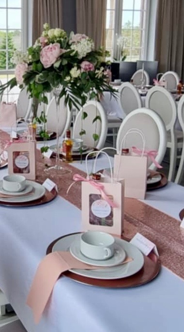 Elegancko nakryty stół na uroczystość: białe obrusy, różowe bieżniki, kwiatowe kompozycje, porcelanowa zastawa, upominki dla gości w pastelowych torebkach z kokardkami.