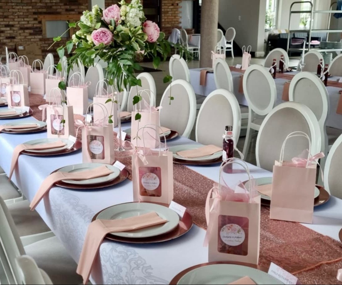 Elegancko nakryty stół z różowymi akcentami: talerze, serwetki, bieżnik z cekinami, upominki dla gości w papierowych torebkach z kokardkami, kwiatowa dekoracja i białe krzesła.
