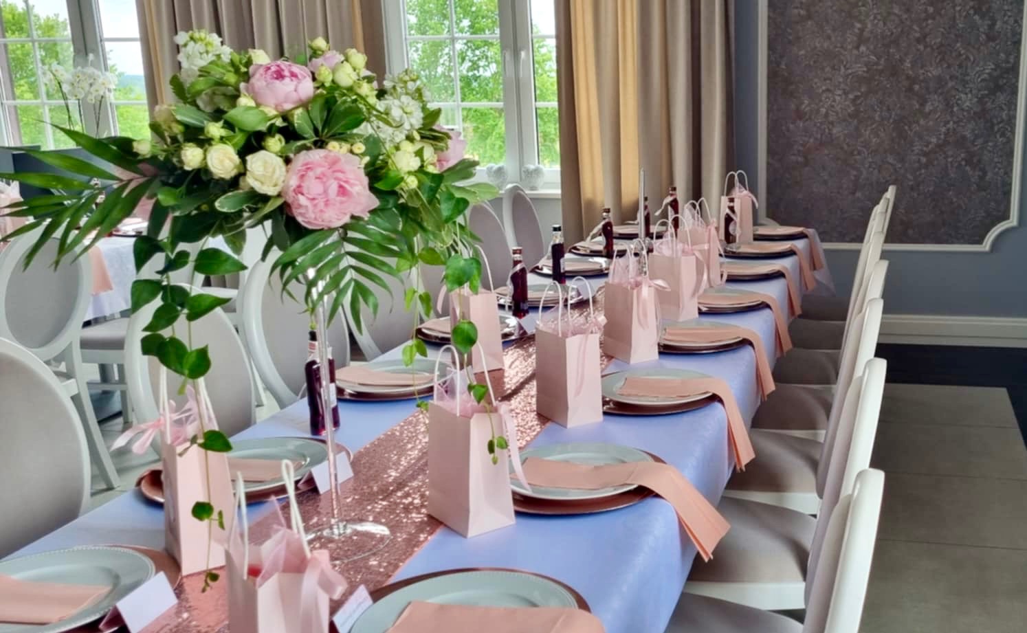 Elegancko nakryty stół weselny z kwiatową dekoracją, różowym bieżnikiem z cekinami, upominkami dla gości w różowych torebkach i butelkami z napojami.