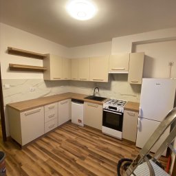 Marcin Jockel Usługi Ogólnobudowlane - Jasna, nowa kuchnia z drewnianymi blatami i panelami podłogowymi. Widoczna drabina malarska sugeruje świeży remont. Uwagę zwracają półki na ścianie.