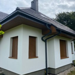 Marcin Jockel Usługi Ogólnobudowlane - Boczny widok domu po renowacji elewacji, z brązowymi roletami okiennymi i ciemną dachówką, na tle pochmurnego nieba i zieleni.