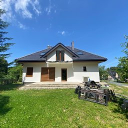 Marcin Jockel Usługi Ogólnobudowlane - Biały dom jednorodzinny z ciemną dachówką i brązowymi okiennicami, otoczony zielonym trawnikiem i drzewami, w słoneczny dzień. Na trawniku leżą elementy konstrukcyjne.