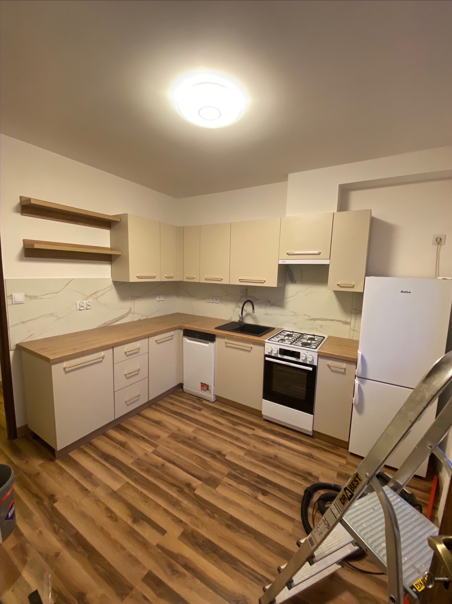 Jasna, nowa kuchnia z drewnianymi blatami i panelami podłogowymi. Widoczna drabina malarska sugeruje świeży remont. Uwagę zwracają półki na ścianie.