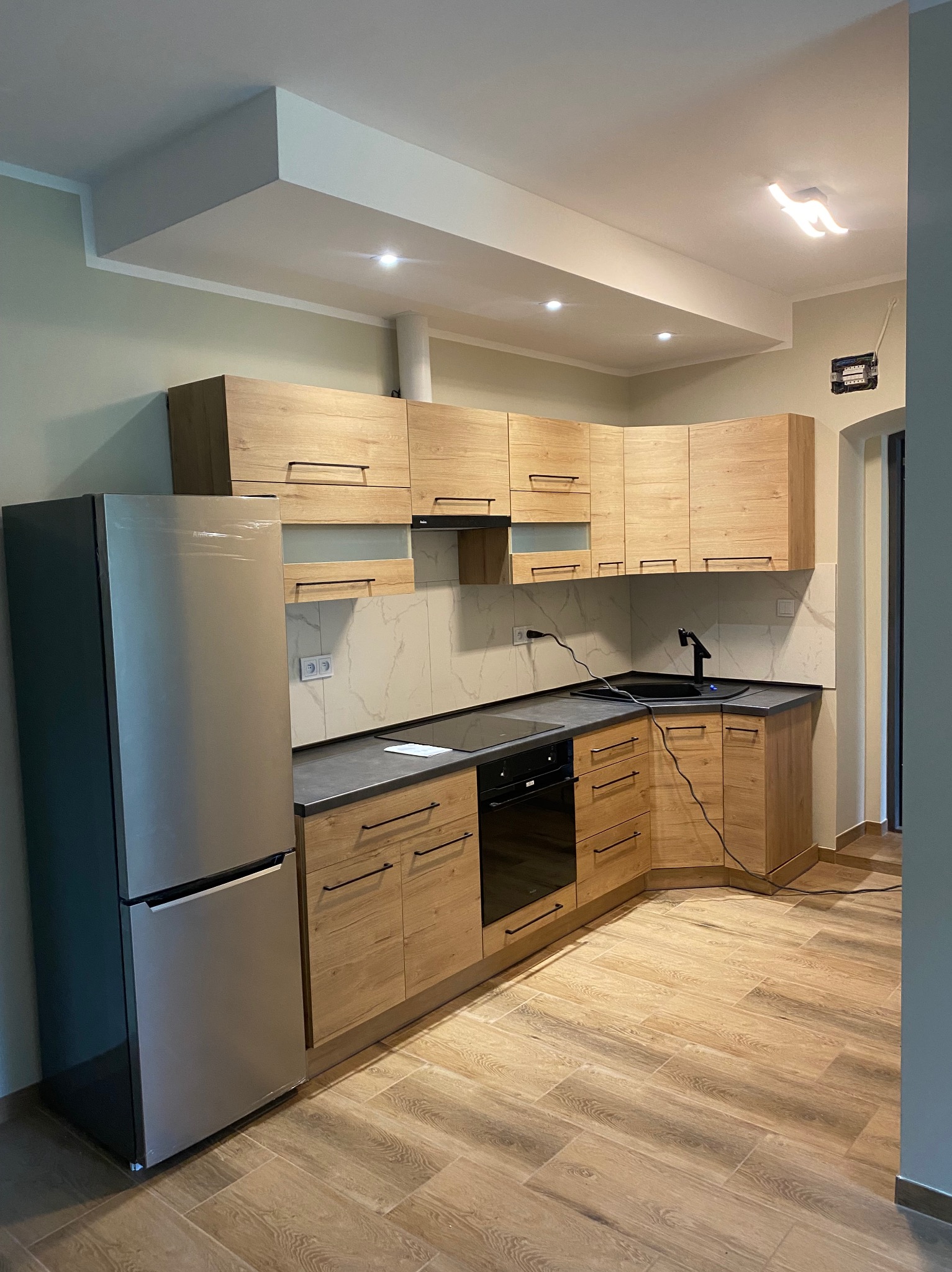 Jasna kuchnia z drewnianymi szafkami, czarnym blatem i sprzętem AGD. Podłoga wyłożona płytkami imitującymi drewno. Nowoczesne oświetlenie sufitowe.